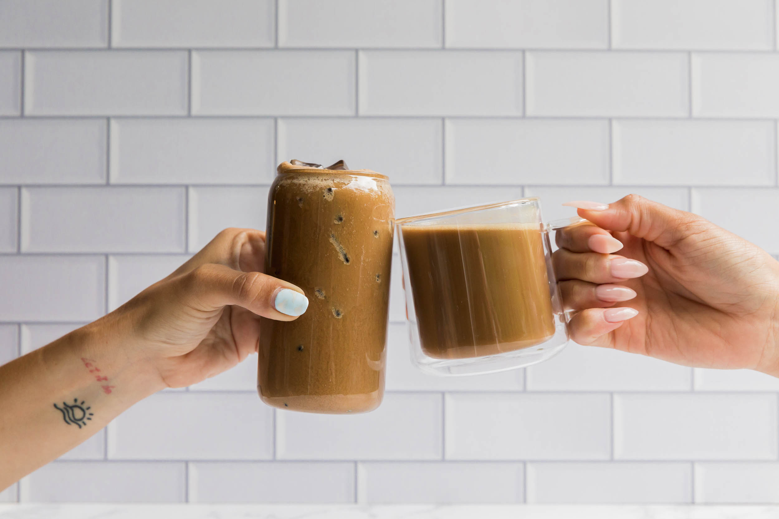 One hand coming in from the left side of the picture holding an iced coffee in a glass which is cheering with a mug filled with hot coffee being held by a hand coming from the right side of the photo with a white tiled background.